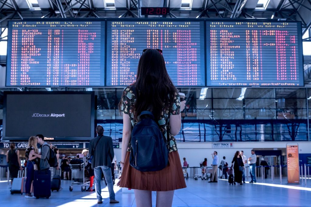 Girl standing back facing the camera in an airport toward the flight information display system