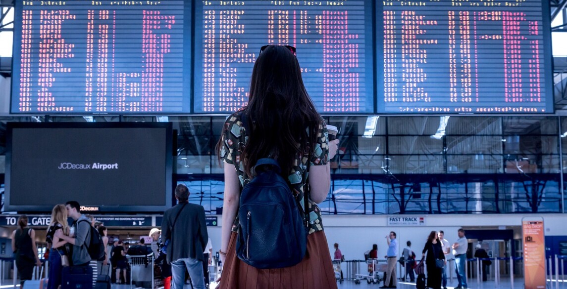 Girl standing back facing the camera in an airport toward the flight information display system