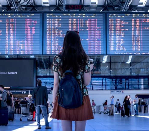 Girl standing back facing the camera in an airport toward the flight information display system