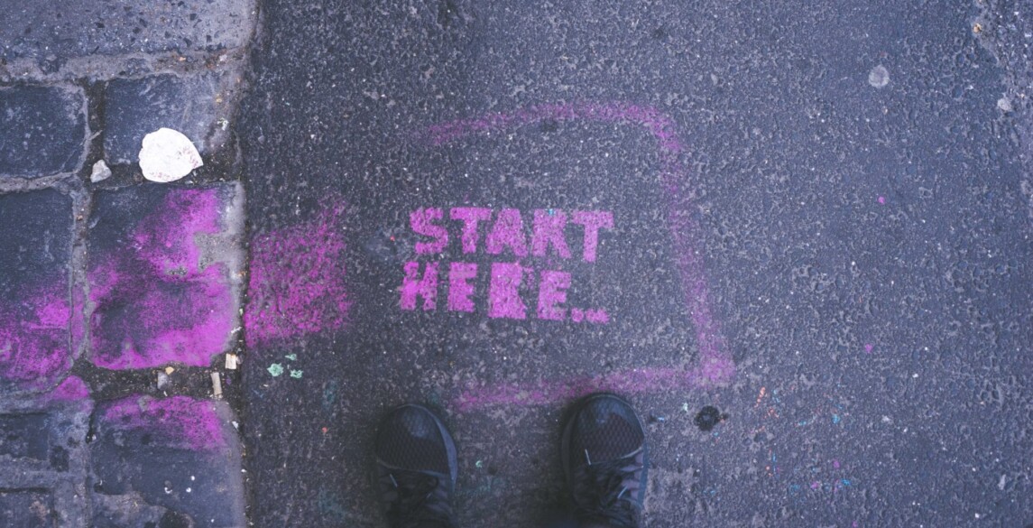 "Start here" spraypainted in pink on the road with a person's feet at the "start here" wording