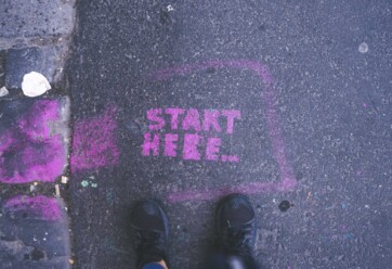 "Start here" spraypainted in pink on the road with a person's feet at the "start here" wording