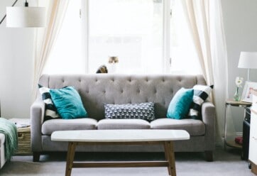 Picture of a living room with a gray couch and teal pillows, against a window.