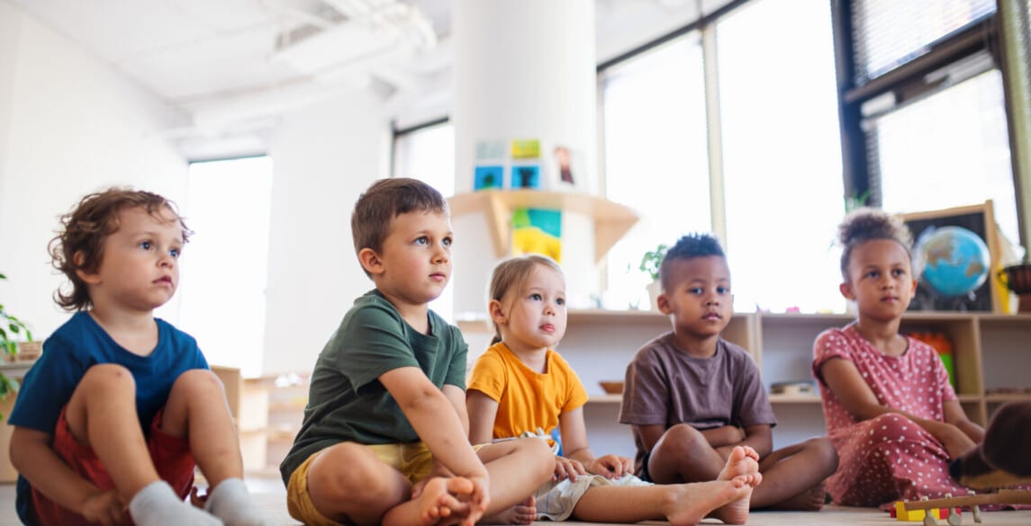 A group of small nursery school children sitting on floor indoors in classroom.