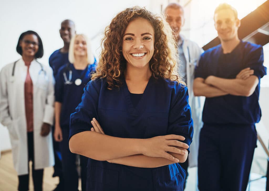 Young female doctor smiling while standing in a hospital corridor with a diverse group of staff in the background.