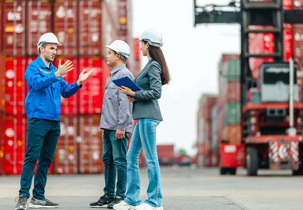 Manager supervising and discussing logistics with two male workers at shipping container yard. Stacked containers and industrial equipment are in the background.