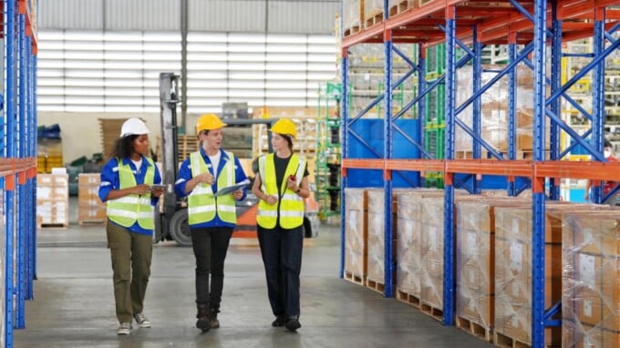 Three individuals walking through a warehouse wearing reflective vests.
